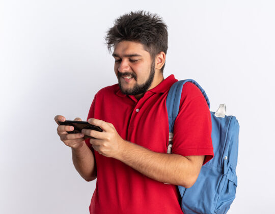 男孩年轻的留着胡子的学生 穿着红色马球衫 背着背包 用智能手机玩游戏 快乐地站着移动学生游戏