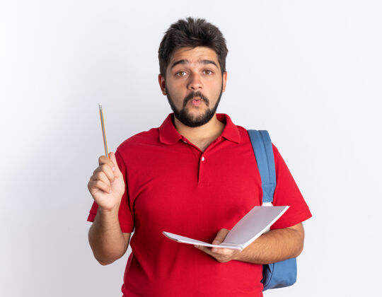 马球一个留着胡子的年轻学生 穿着红色马球衫 背着背包 手里拿着笔记本和铅笔 站在那里显得很惊讶红色小伙子惊喜