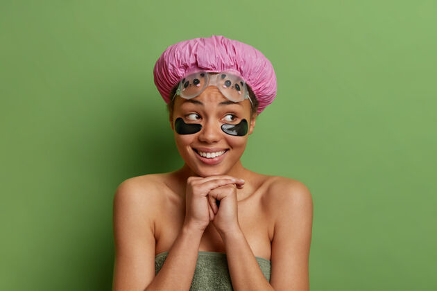 治疗快乐的年轻非洲裔美国妇女保持双手放在下巴下面的水平镜头微笑着轻轻地在眼睛下面用浴巾包裹的美容垫隔离在绿色的墙上年轻下巴满意