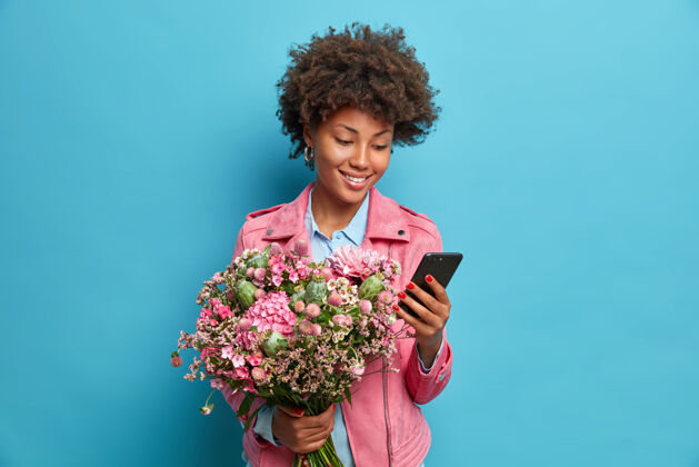 网站积极的年轻女子在她的生日接受祝贺手持手机花束穿着粉色夹克隔着蓝色的墙查看新闻稿女性姿势电话