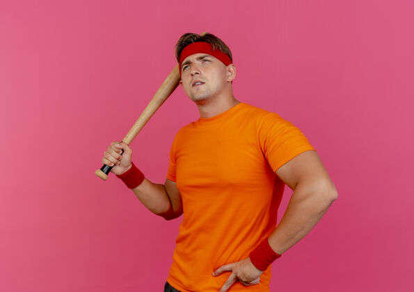 腰体贴的年轻帅气的运动型男人戴着头带和腕带拿着棒球棒 手放在腰上 孤零零地看着粉红色的墙上年轻感觉腕带