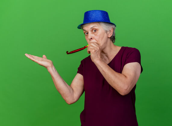 帽子惊讶的老妇人戴着党的帽子吹哨子 并保持在绿色的手打开惊喜手女人