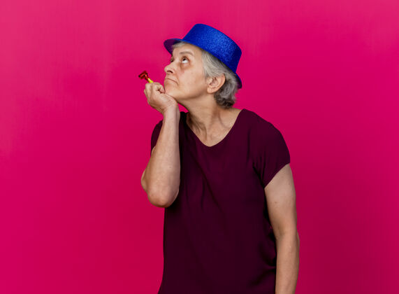 手体贴的老妇人戴着聚会帽 手放在下巴上 手里拿着哨子 抬头看着粉红色的脸帽子哨子聚会