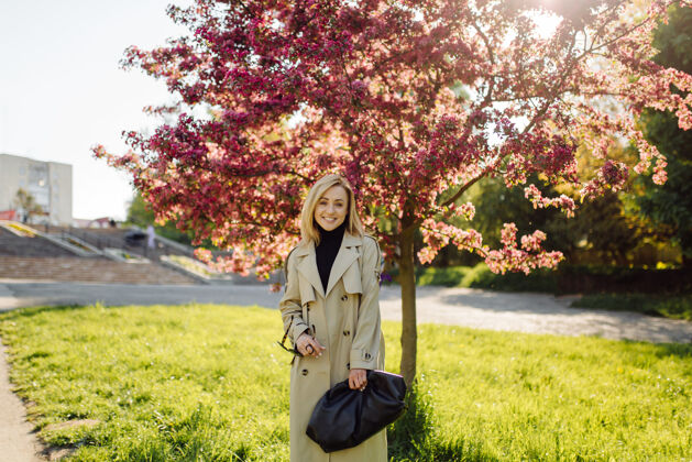 别致在阳光明媚的春日 穿着战壕的白种人金发女人在公园散步 开心地微笑着头发时髦秋天