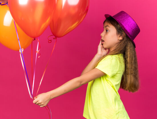 帽子惊讶的小白种女孩 戴着紫色派对帽 手放在脸上 看着粉色墙上的氦气球 还有复制空间小气球白种人