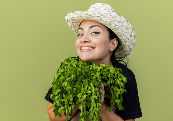 微笑年轻漂亮的女园丁 围着围裙 戴着帽子 手里拿着新鲜的药草 站在浅绿的墙上 面带微笑地看着前面美丽花园感觉