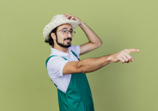 人年轻的留着胡须的园丁 穿着连体衣 戴着帽子 食指指着站在浅绿色墙上的东西指点人人