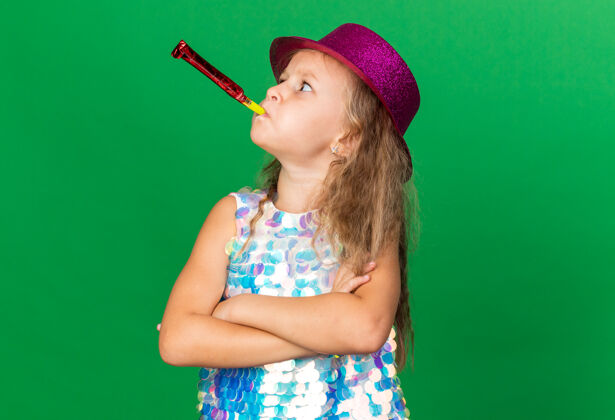 吹恼怒的金发小女孩 戴着紫色的派对帽 双臂交叉站着 吹着派对的哨子 看着绿色墙壁上孤立的一面 还有复制空间生日胳膊小