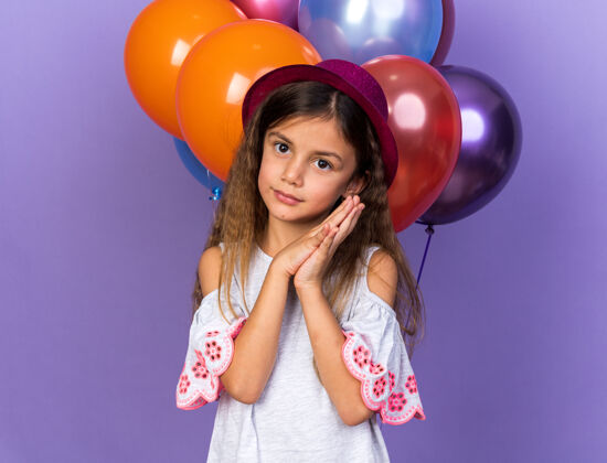 手戴着紫色派对帽的高加索小女孩手牵着手站在氦气球前 氦气球被隔离在紫色的墙上 留着复制空间生日气球抱