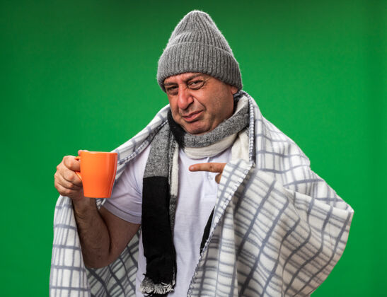 男人未租成年病高加索男子脖子上围着围巾 戴着格子布包裹的冬季帽子 手持并指着隔离在绿色墙壁上的杯子 留有复印空间指着围巾疾病
