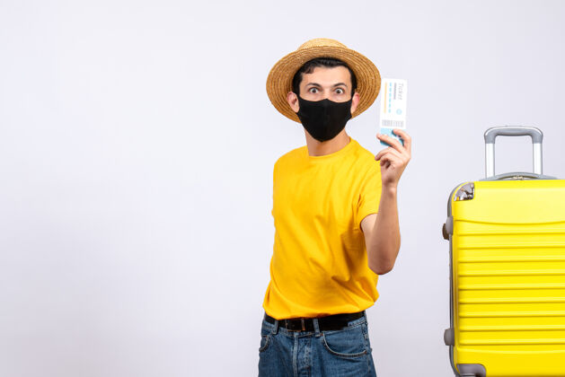 视图正面图穿着黄色t恤的游客站在黄色手提箱旁举着旅行票前面向上帽子