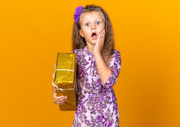 脸震惊的小金发女孩把手放在脸上 拿着礼品盒隔离在橙色墙上的复制空间抱着女孩手