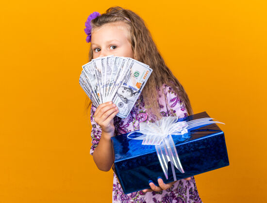 小个子高兴的小金发女孩拿着礼品盒和钱在她面前孤立的橙色墙上复制空间钱孤立的抱着