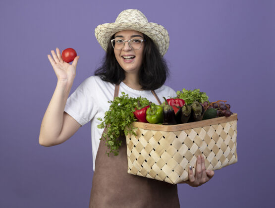 眼镜戴着眼镜 身着制服 头戴园艺帽的快乐的年轻黑发女园丁把菜篮子和西红柿隔离在紫色的墙上园艺西红柿年轻人