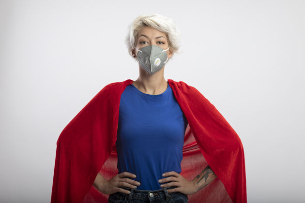 手自信的女超人身穿红色斗篷 戴着医用面罩 双手放在隔离在白墙上的腰上人自信腰