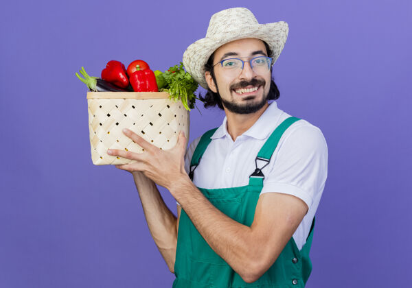 衣服年轻的留着胡须的园丁 穿着连体衣 戴着帽子 拿着装满蔬菜的箱子 站在蓝色的墙上 面带微笑地看着前面连身衣市民人