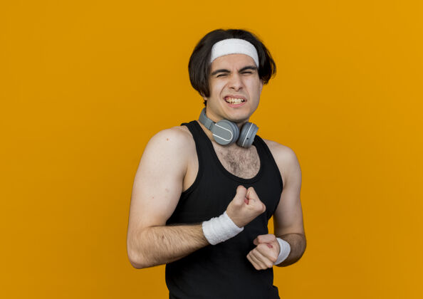 头带穿着运动服 头戴耳机 脖子上戴着头带的年轻人站在橘色的墙上看着紧握的拳头姿势头