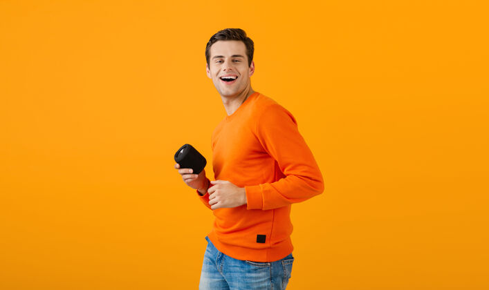 时髦穿着橙色毛衣的时髦微笑的年轻人拿着无线扬声器快乐地听音乐享受橙色的乐趣男性扬声器人物