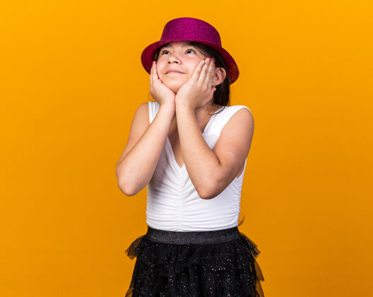 手戴着紫色派对帽的年轻白种女孩高兴地把手放在脸上 孤立地仰望着橘黄色的墙上 还有复制空间白种人年轻女孩