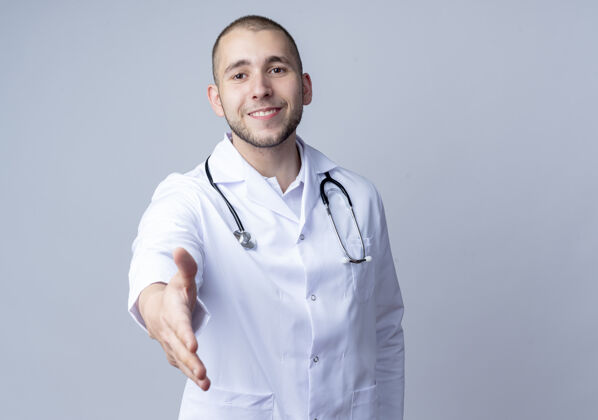 人面带微笑的年轻男医生 身穿医用长袍 脖子上戴着听诊器 在前面伸出一只手 在白色的墙上打招呼伸展衣服听诊器