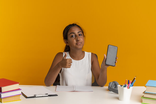 背景看着照相机 年轻的女学生正坐在书桌旁 手里拿着学习工具 举着大拇指向上书桌工具