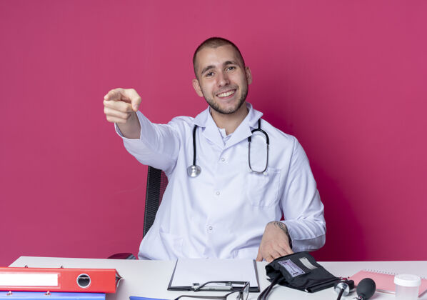 医生面带微笑的年轻男医生穿着医用长袍 听诊器坐在办公桌旁 工作工具笔直地指向粉红色的墙上工具桌子人