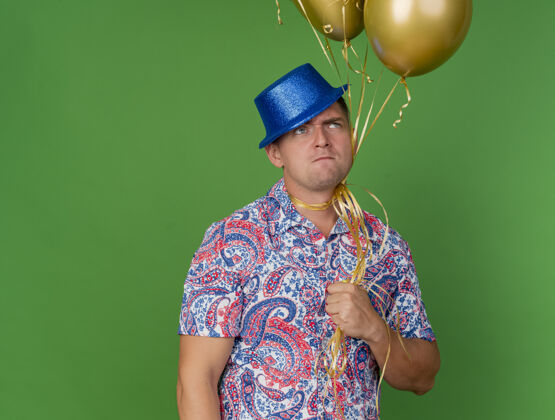 围着一个体贴的年轻人戴着蓝色的帽子 脖子上绑着气球 闭着眼睛站在绿色的地面上绿色派对封闭