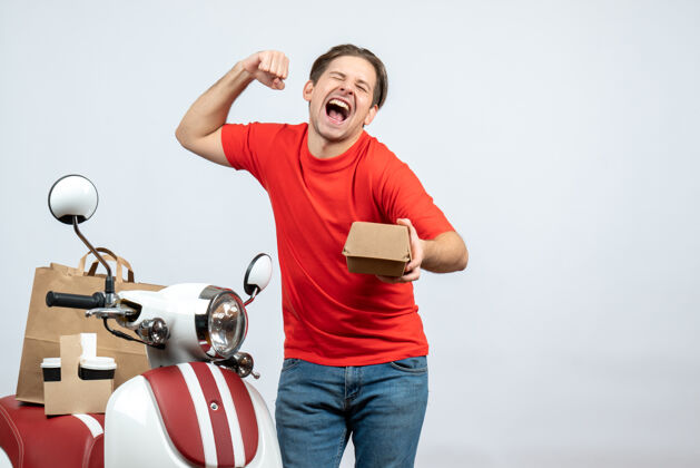 健身骄傲的送货员穿着红色制服站在滑板车旁 手里拿着白色背景的小盒子抱运动盒子