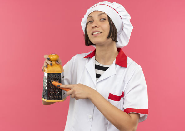 感激面带微笑的年轻女厨师身着厨师制服 在粉红色的墙上用烤炉烤胡萝卜感情姿势人