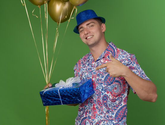 派对面带微笑的年轻人戴着蓝帽子 拿着气球 指着礼品盒孤立地站在绿色的气球帽子盒子