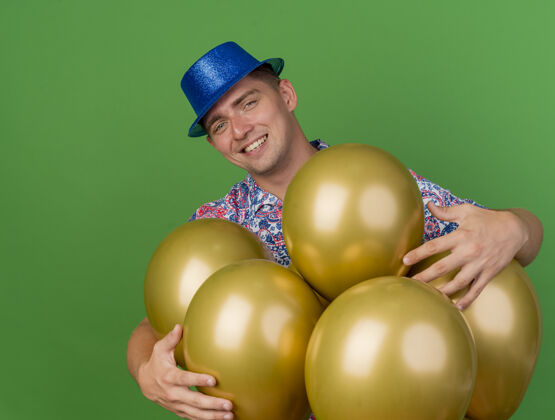 站着微笑着的年轻人戴着蓝帽子站在绿色的气球后面气球绿色小伙子