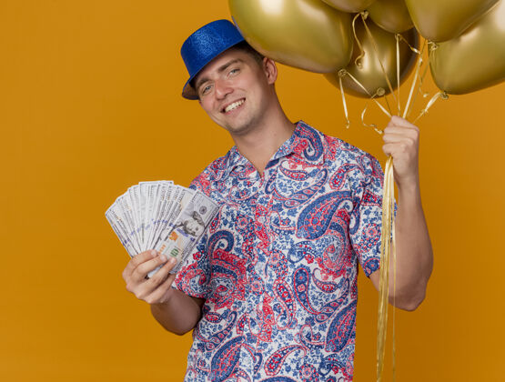 蓝色带着微笑的年轻人戴着蓝色的帽子拿着气球 把钱孤立在橙色上帽子派对年轻人