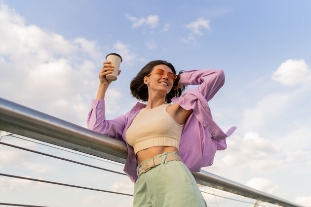 休闲幸福的女人 完美的身材 穿着时尚的紫色超大号衬衫 一边在摩登桥上享受着一杯咖啡时尚微笑潮流