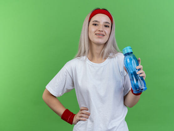 水面带微笑的年轻运动女性戴着背带 戴着头带和腕带 拿着水瓶 把手放在隔离在绿色墙上的腰上持有运动穿