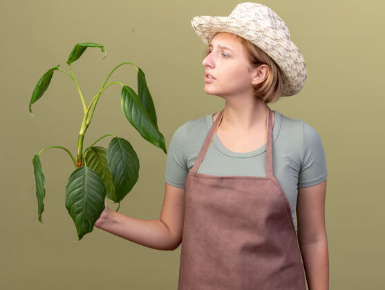 帽子不知所措的年轻斯拉夫女园丁戴着园艺帽拿着橄榄绿上的植物看着无知园艺女性