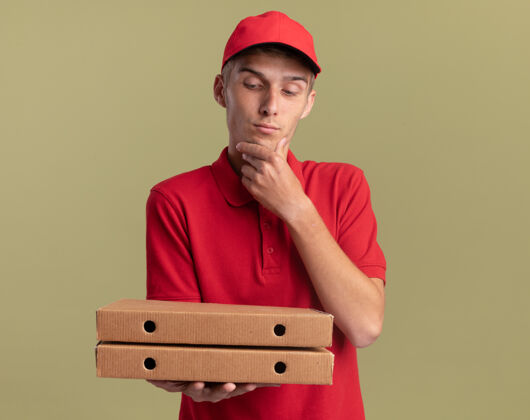 金发年轻的金发送货员高兴地把手放在下巴上拿着 看着橄榄绿的比萨饼盒下巴送货男孩