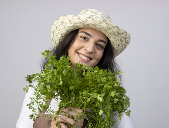 园艺微笑的年轻黑发女园丁穿着制服 戴着园艺帽 把香菜孤立地放在白色墙壁上 留有复制空间持有花园制服