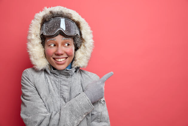 护目镜冬天的乐趣和滑雪的概念高兴的深色皮肤的非裔美国妇女在外套指向远处的空白空间显示方向 以度假胜地享受十二月的季节指向快乐灰色