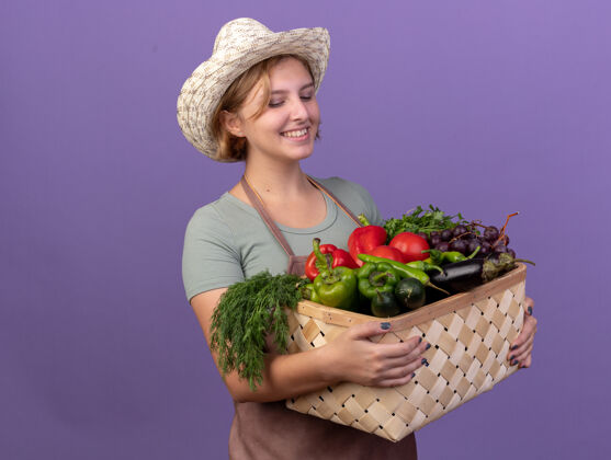 斯拉夫人高兴的年轻斯拉夫女园丁戴着园艺帽 拿着紫色的菜篮看着篮子花园蔬菜