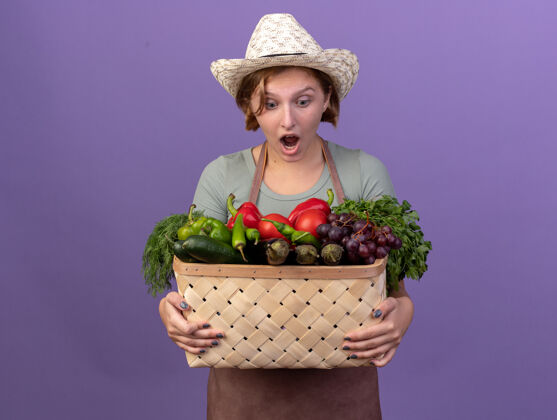 斯拉夫兴奋的年轻斯拉夫女园丁戴着园艺帽 拿着紫色的菜篮看着帽子蔬菜篮子