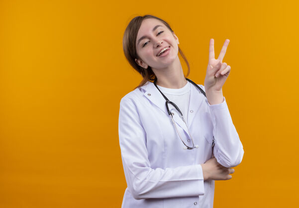 长袍快乐的年轻女医生穿着医用长袍和听诊器在隔离的橙色空间和复印空间做和平标志医疗听诊器医生