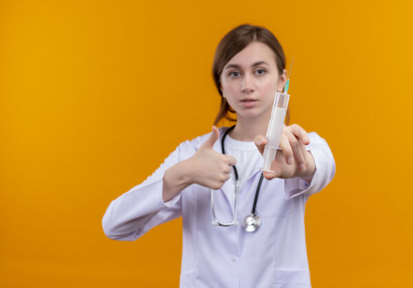注射器一位面容严肃的年轻女医生 穿着医用长袍 戴着听诊器 伸展着注射器 在有复印空间的孤立橙色空间上竖起大拇指医生太空向上