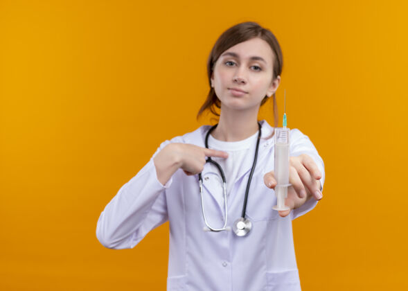 听诊器一位面容严肃的年轻女医生 穿着医用长袍 手持听诊器 指着自己 将注射器伸向一个孤立的橙色空间和复印空间她自己穿指向