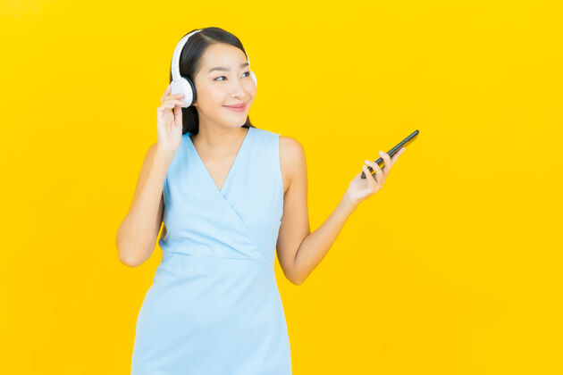 成人画像美丽的亚洲年轻女子戴着耳机和智能手机在黄色的墙上听音乐女性年轻脸
