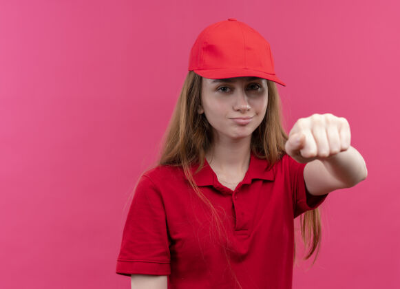 传递穿着红色制服的自信的年轻送货女孩在粉色的空间里伸出她的拳头制服自信伸展