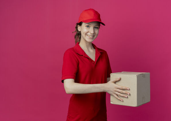 背景微笑着的年轻漂亮的送货女孩 穿着红色制服 戴着帽子 拿着纸箱 在深红色的背景上与复印空间隔离开来深红色制服盒子