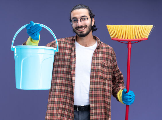 表情微笑的年轻帅气的清洁工 穿着t恤 戴着手套 拿着桶 把拖把隔离在蓝色的墙上帅哥男人拖把