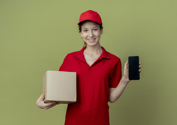 漂亮微笑着的年轻漂亮的送货女孩 穿着红色制服 戴着帽子 展示着手机 拿着橄榄绿背景上孤立的纸箱背景年轻持有