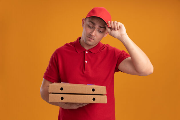 公民年轻的送货员穿着制服 戴着帽子 看着隔离在橙色墙上的比萨饼盒人盒子披萨