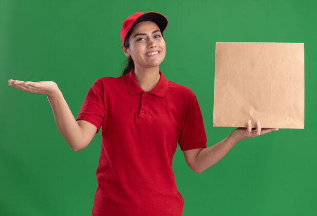 纸带着微笑的年轻送货女孩穿着制服 戴着帽子 手里拿着纸制食品包 手被隔离在绿色的墙上微笑帽子制服